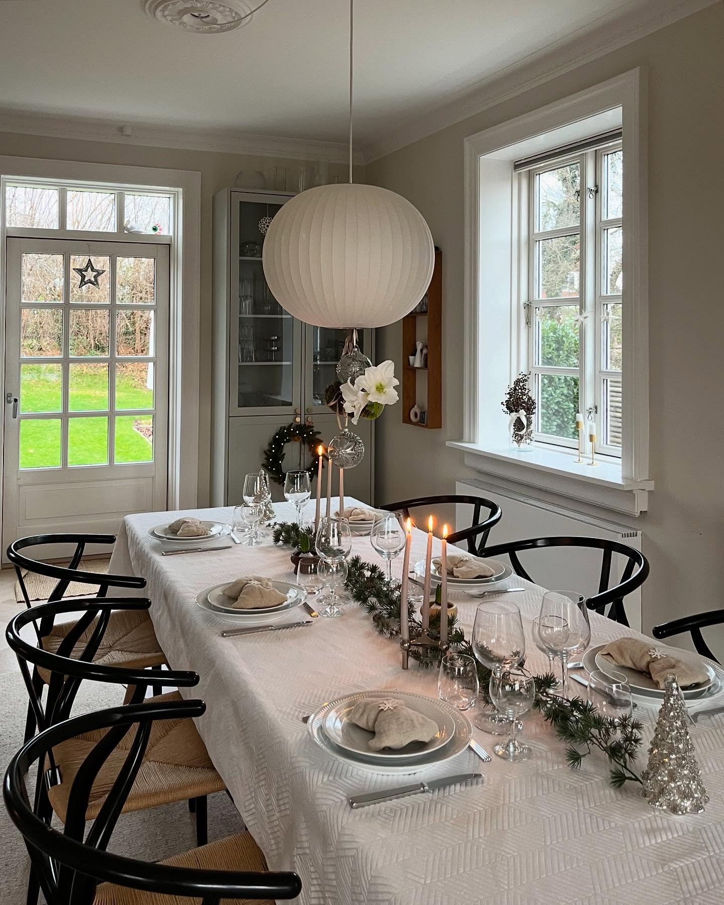 A bright dining room with a table covered in a white textured tablecloth, set with layered clear glass and white plates, light beige cloth napkins, and a simple garland of evergreen branches down the center.