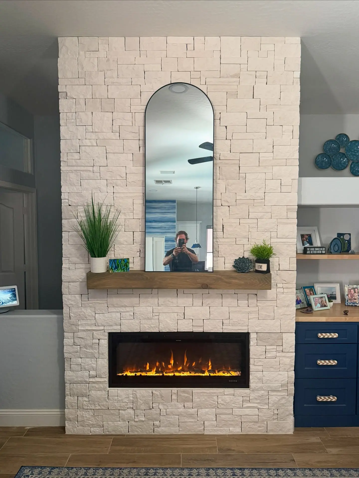 Wide, horizontal electric fireplace set into a floor-to-ceiling accent wall clad in stacked white stone, topped by a rustic wood floating mantel and a tall arched mirror.