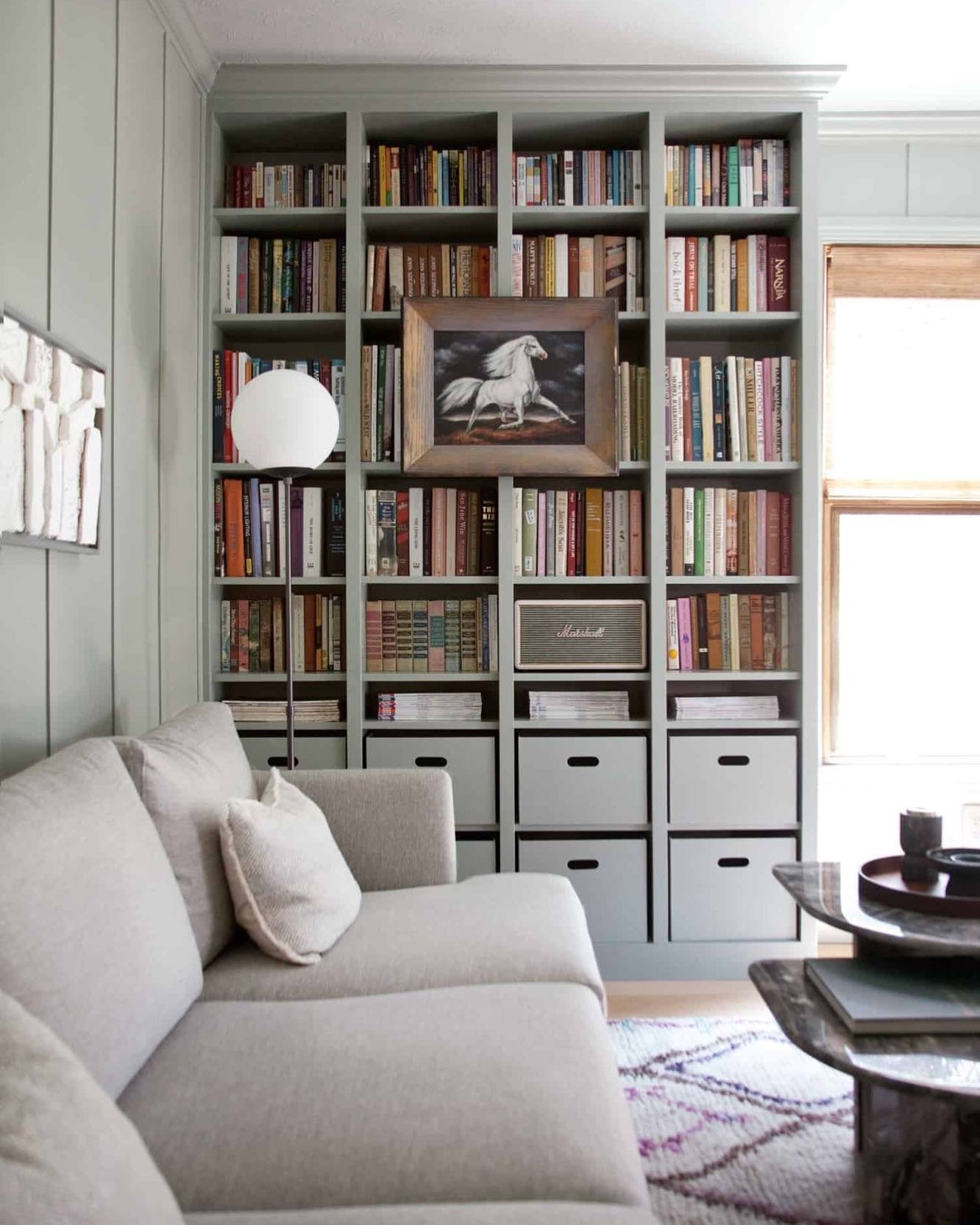 Floor-to-ceiling built-in shelving painted a soft sage green, combining open book compartments with enclosed drawers at the bottom, centered with a framed horse painting.