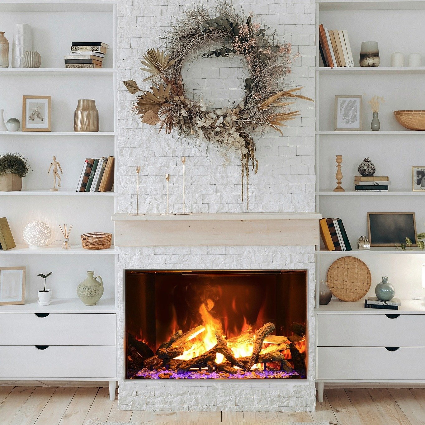 Wide electric fireplace set into a white painted brick or textured tile chimney breast, flanked by white built-in bookcases and cabinets, featuring a large dried floral wreath above.