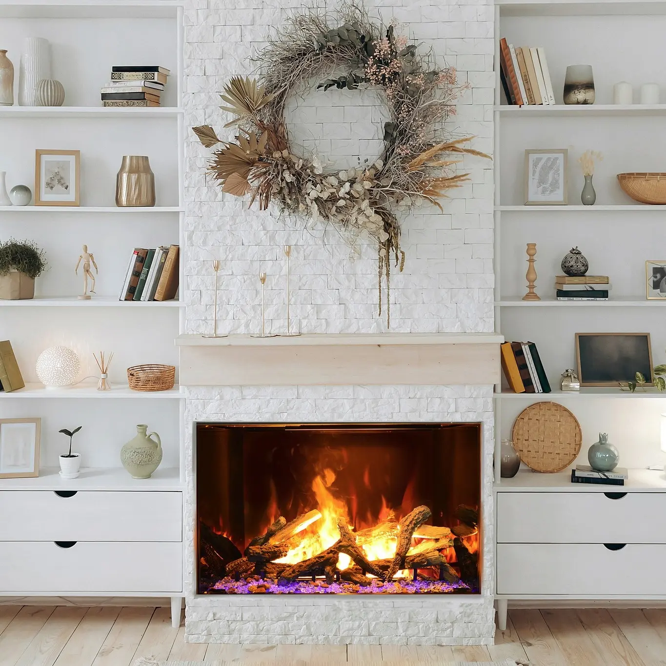 Wide electric fireplace set into a white painted brick or textured tile chimney breast, flanked by white built-in bookcases and cabinets, featuring a large dried floral wreath above.