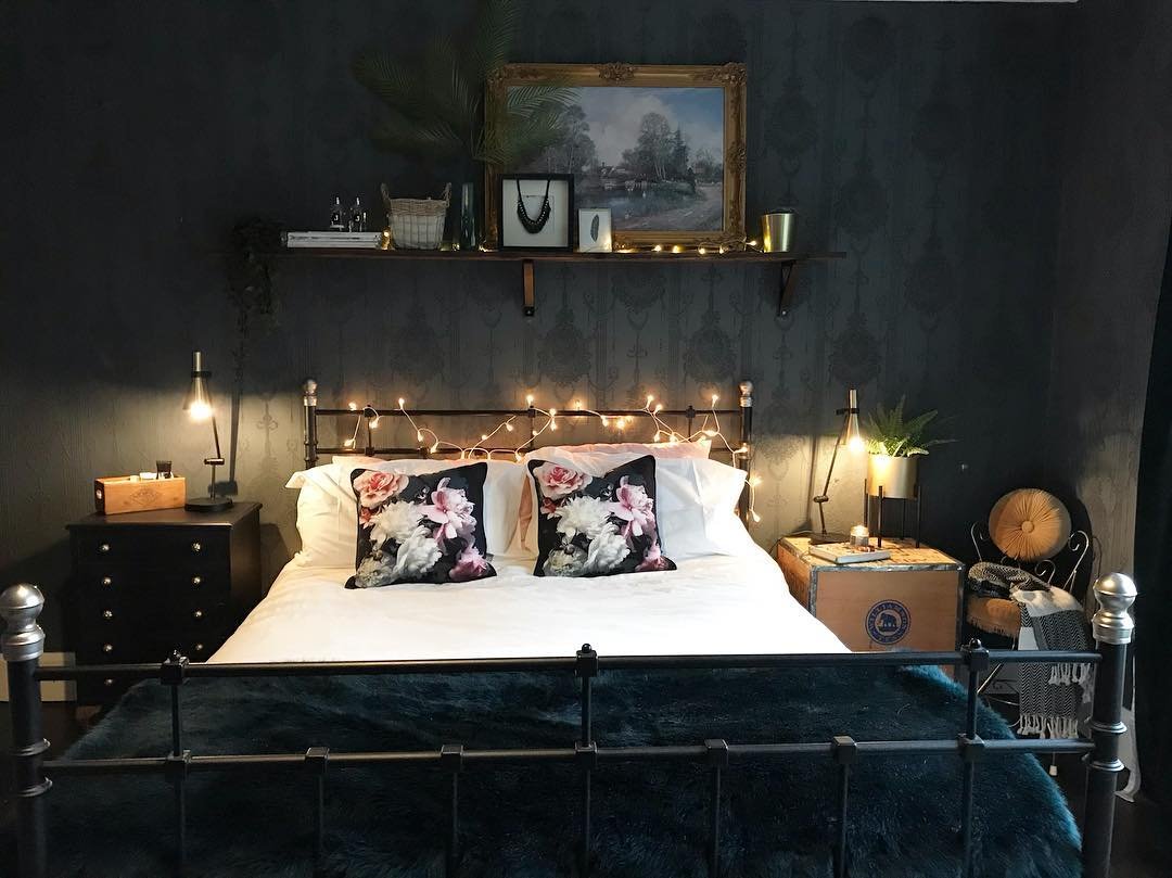 Dark bedroom with textured black damask wallpaper, a black metal frame bed decorated with fairy lights, and contrasting white bedding and floral pillows.