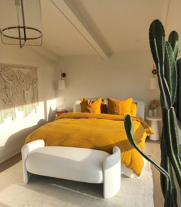 A bright, modern bedroom with a low white bench at the foot of the bed, which is covered in vibrant mustard yellow linen. A tall cactus stands in the foreground.