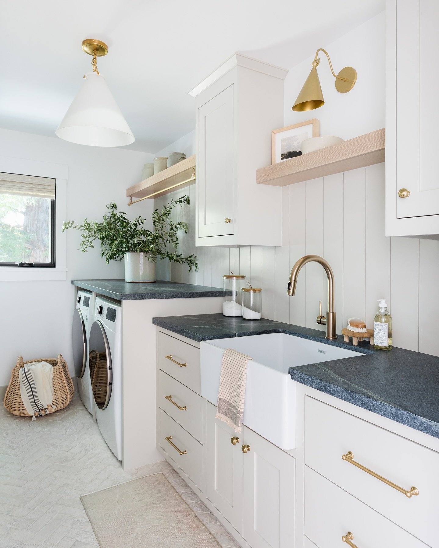 A bright white and gold laundry room featuring dark blue/gray countertops, a white apron-front sink, light wood floating shelves, and a washer and dryer tucked beneath a long counter.