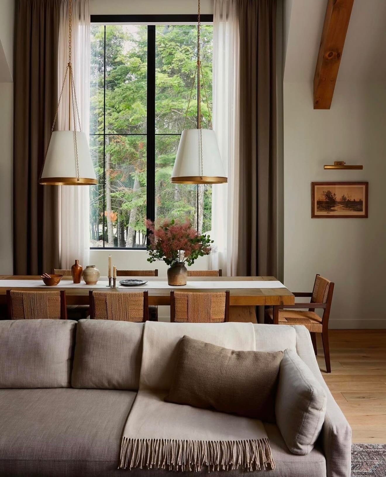 A long wooden dining table set in front of a tall window overlooking a forest, flanked by brown curtains and two large white cone-shaped pendant lights.