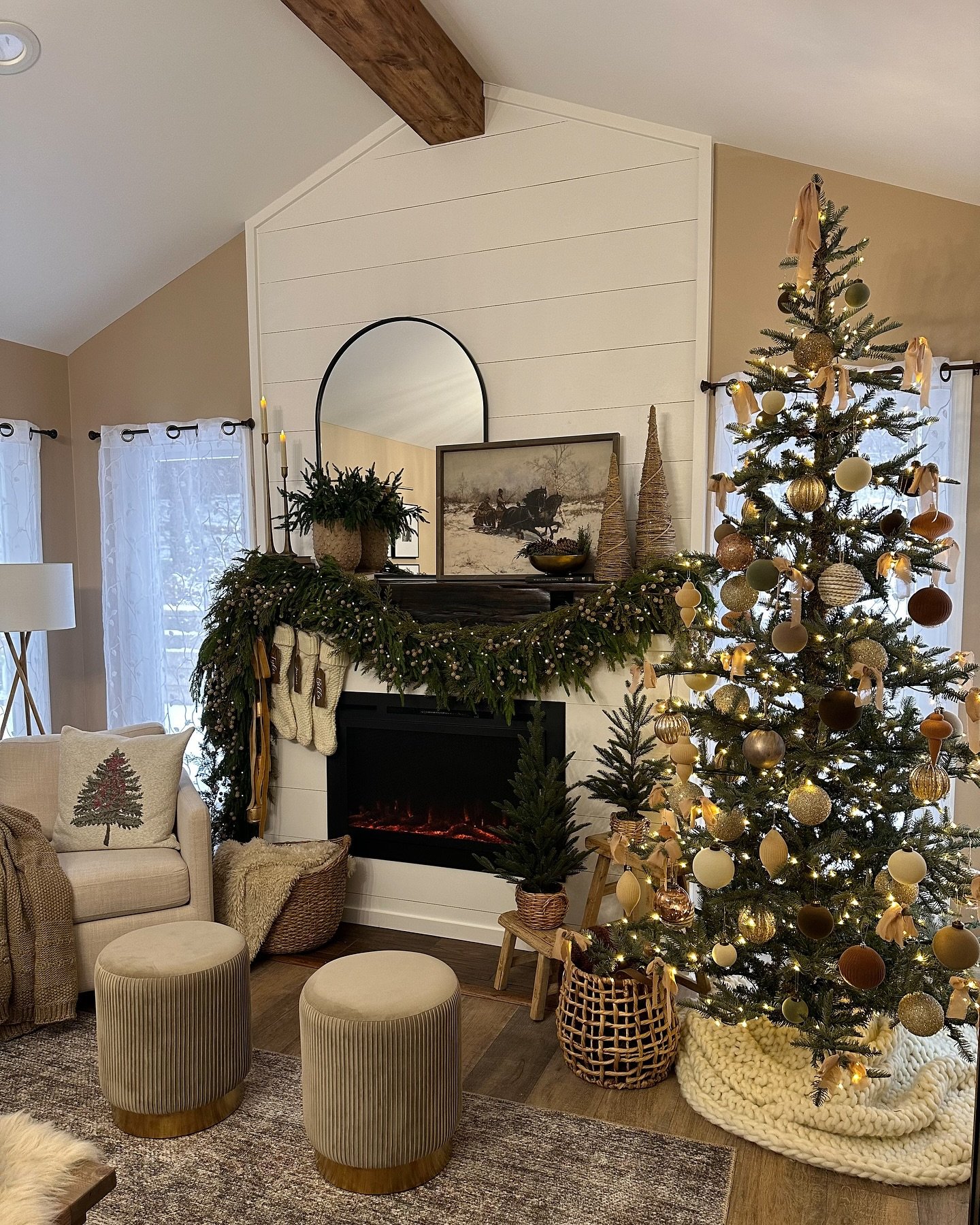 Fireplace mantel with white shiplap above, decorated with thick pine garland, tan stockings, wicker trees, and a framed winter landscape painting.