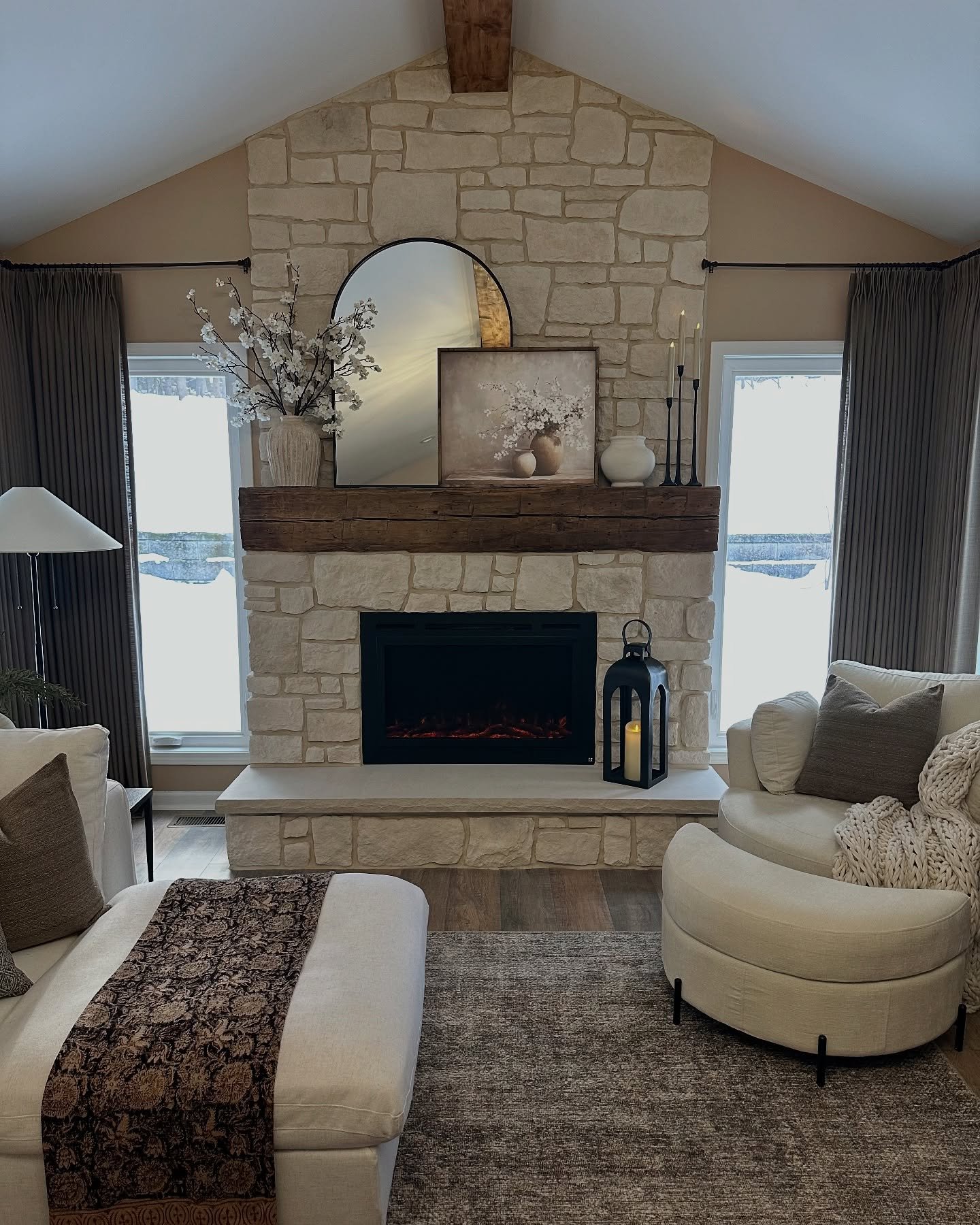 Vaulted living room with a floor-to-ceiling cream stone fireplace, thick distressed wood mantel, round mirror, and curved seating.