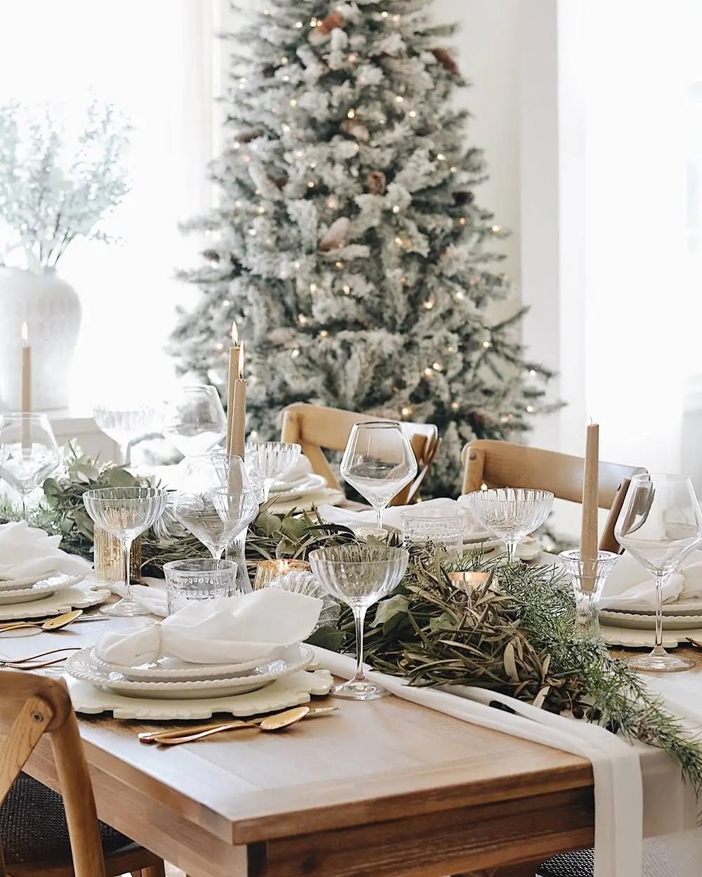 A bright Christmas dining table with a light wood surface, set with a sheer white runner, layered white plates, gold flatware, and a lush centerpiece of olive branches, rosemary, and eucalyptus.