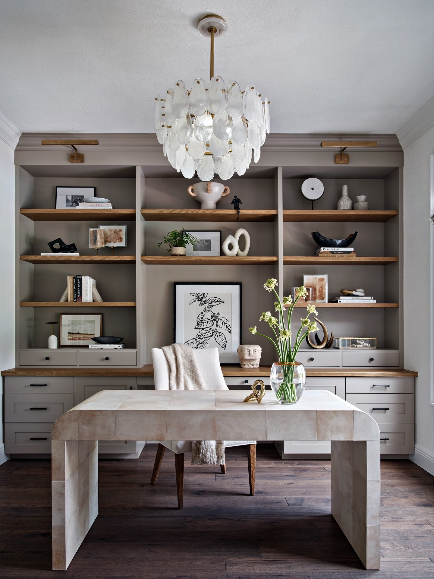 Luxurious home office with greige built-in shelves, a light stone waterfall desk, a plush white chair, and a striking feather or capiz shell chandelier.