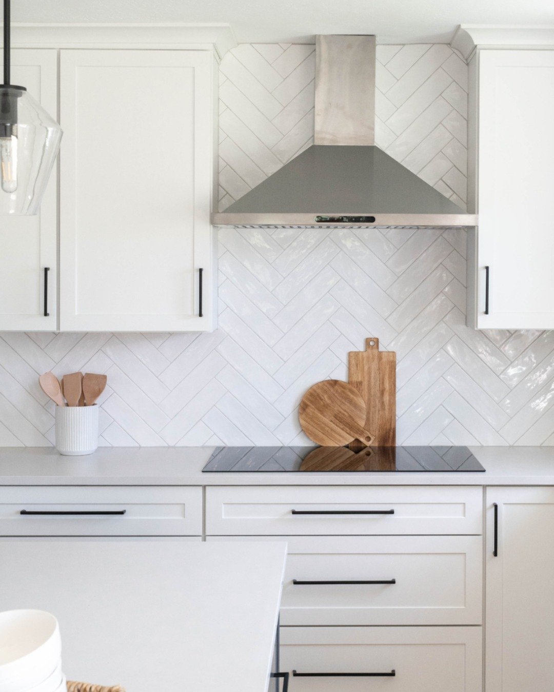 All-white modern kitchen featuring a glossy white subway tile in a herringbone pattern backsplash, white shaker cabinets with black hardware, and a stainless steel hood.