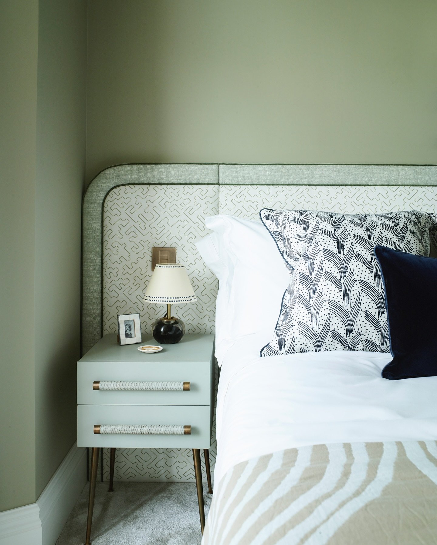 Wide headboard panel with rounded corners upholstered in a light grey fabric with a maze-like pattern, framed by solid sage green piping against a matching wall.