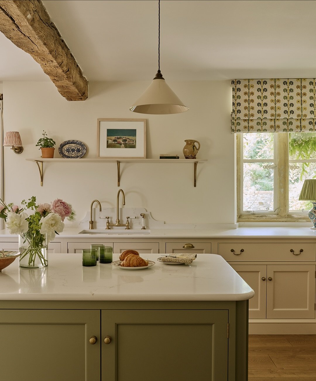 Neutral kitchen with a sage green island and cream cabinets, featuring an open window dressed with a rolled-up Roman blind in a small yellow floral print fabric.