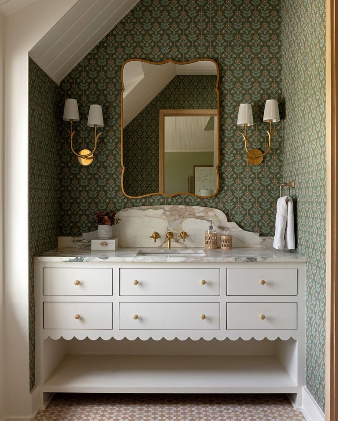 Powder room with green floral wallpaper, a white vanity featuring a scalloped apron, and a brass-framed mirror.