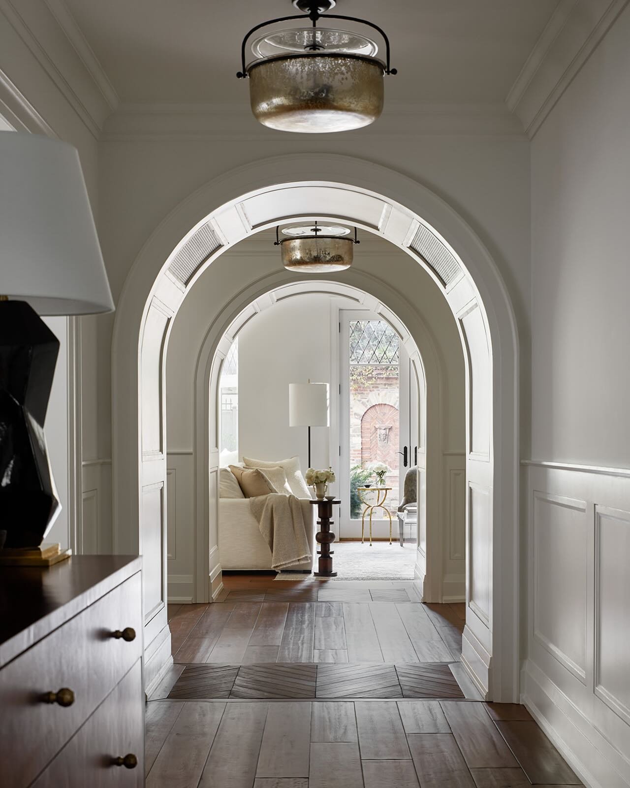 Elegant hallway with white paneling and a dark wood floor, featuring a series of arched openings and antique mercury glass drum pendants.