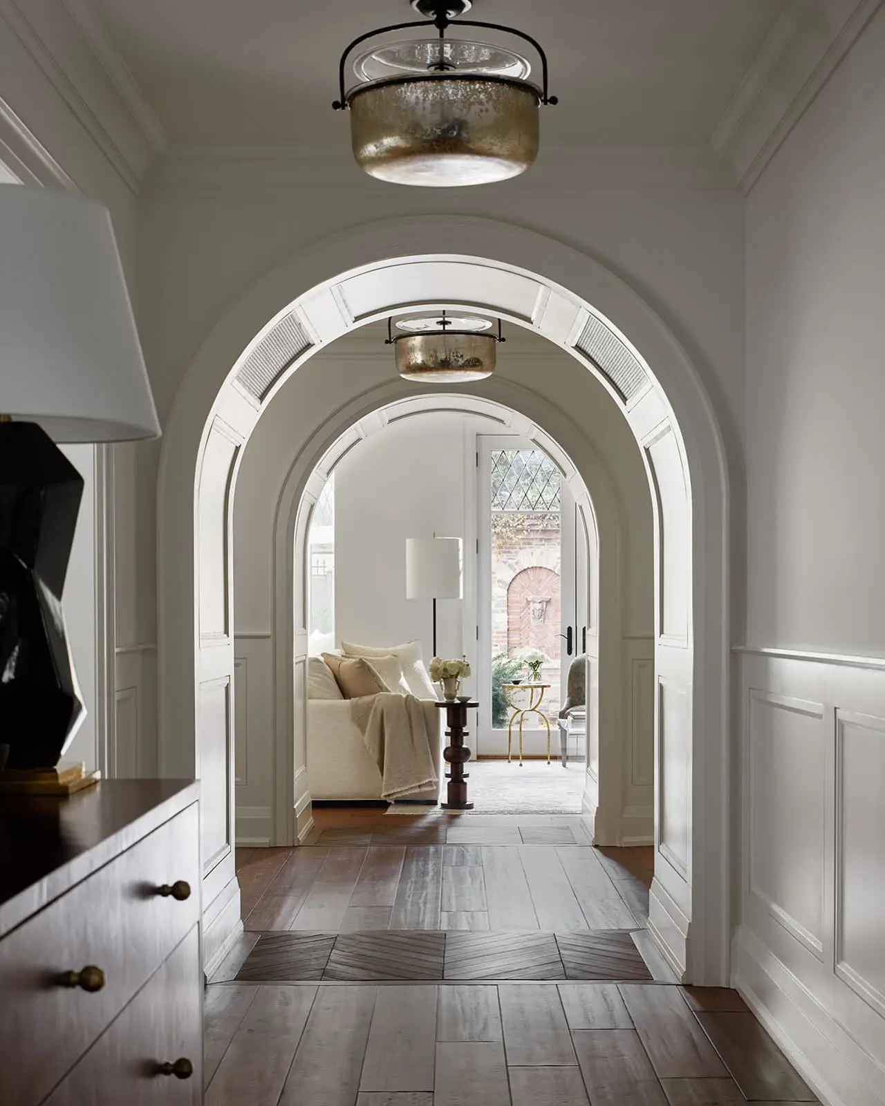 Elegant hallway with white paneling and a dark wood floor, featuring a series of arched openings and antique mercury glass drum pendants.