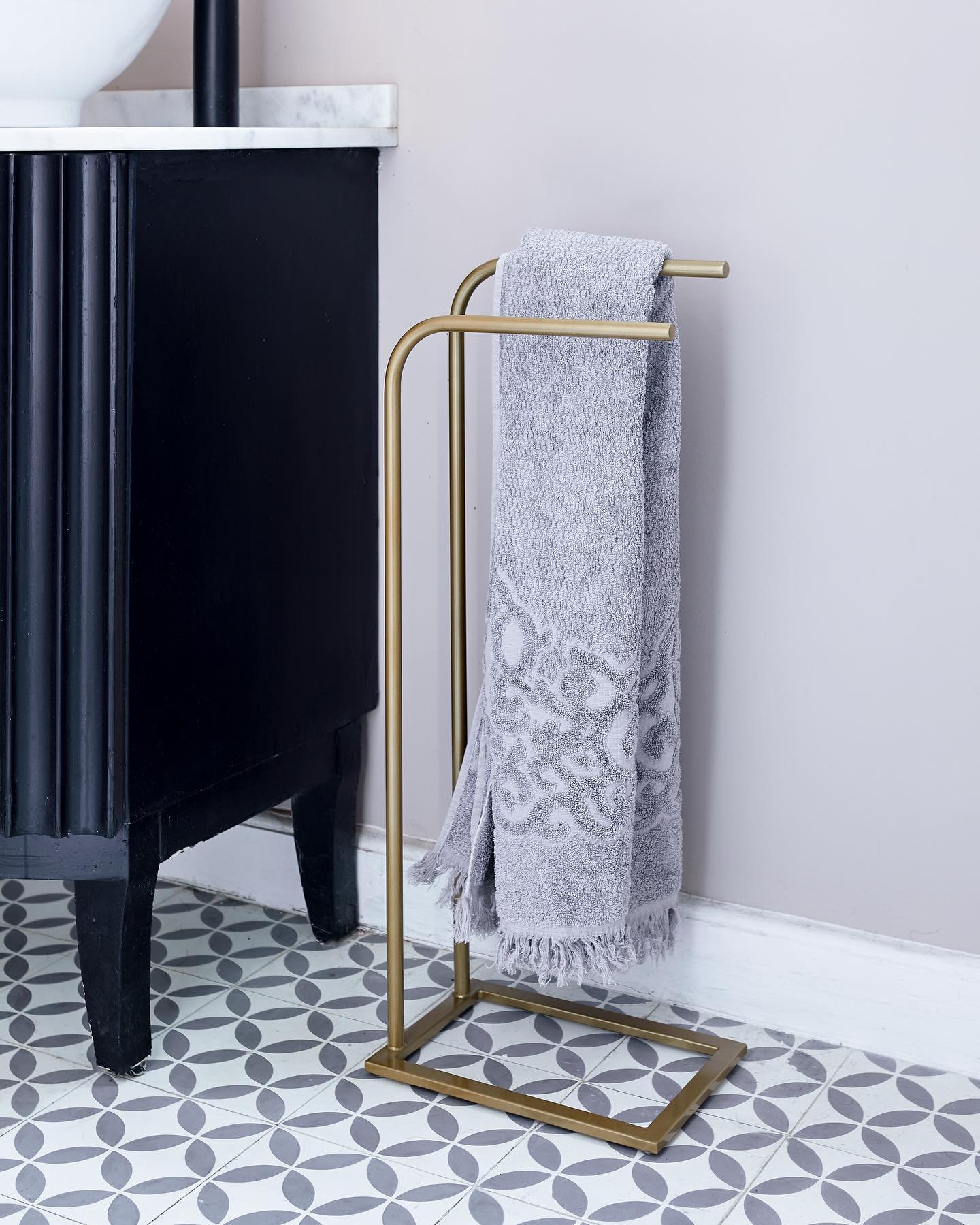 Freestanding brass metal towel stand holding a folded grey hand towel, placed next to a black fluted vanity cabinet on a grey patterned tile floor.