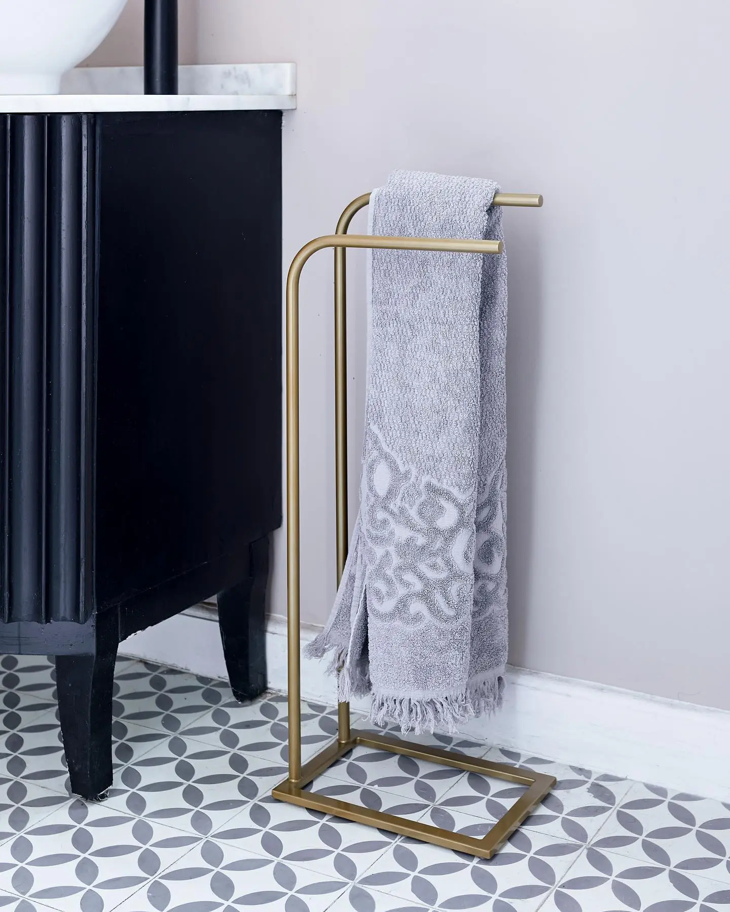 Freestanding brass metal towel stand holding a folded grey hand towel, placed next to a black fluted vanity cabinet on a grey patterned tile floor.