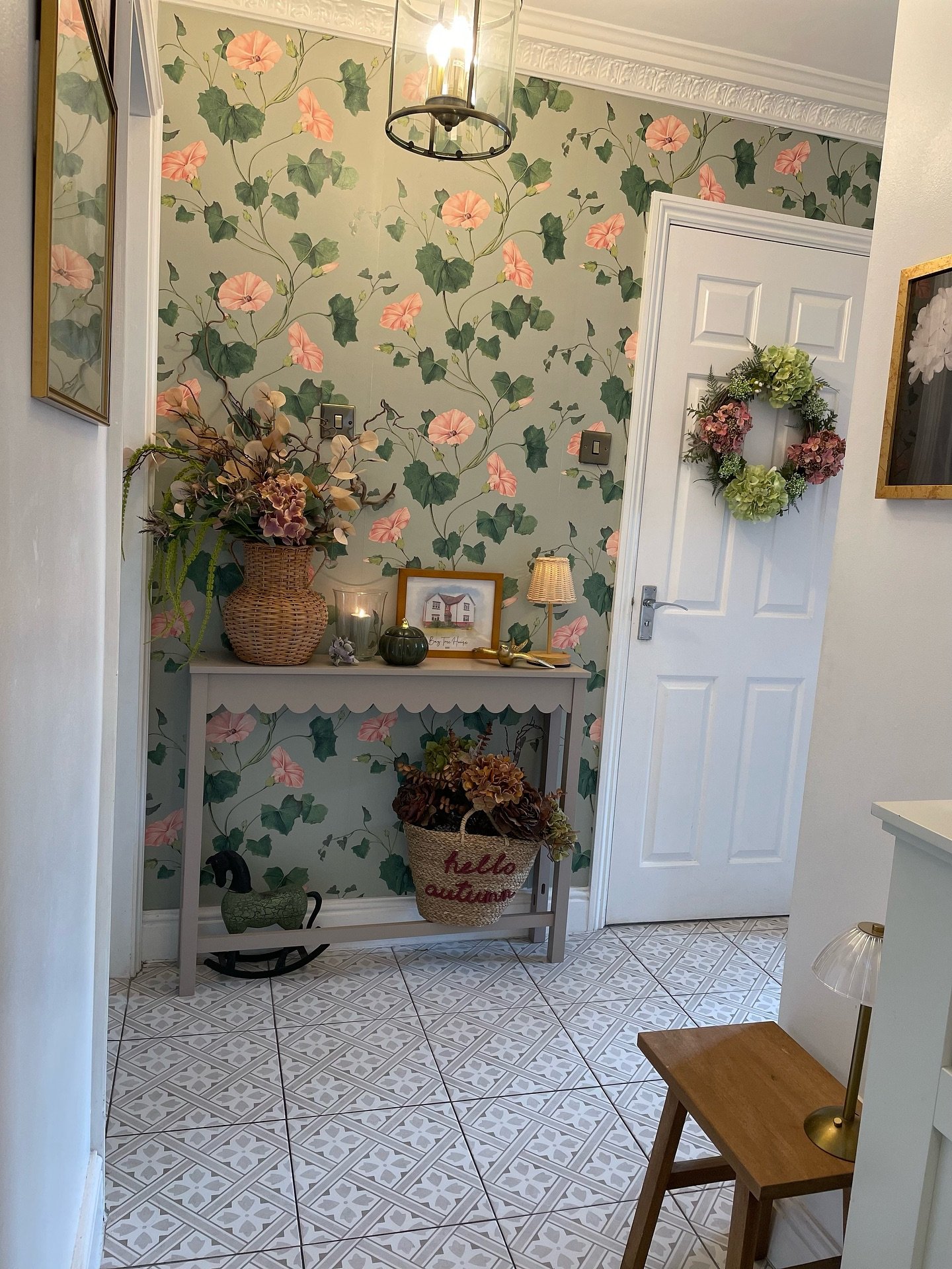 A small entryway with a bold green and pink floral wallpaper a scalloped grey console table and patterned floor tiles.
