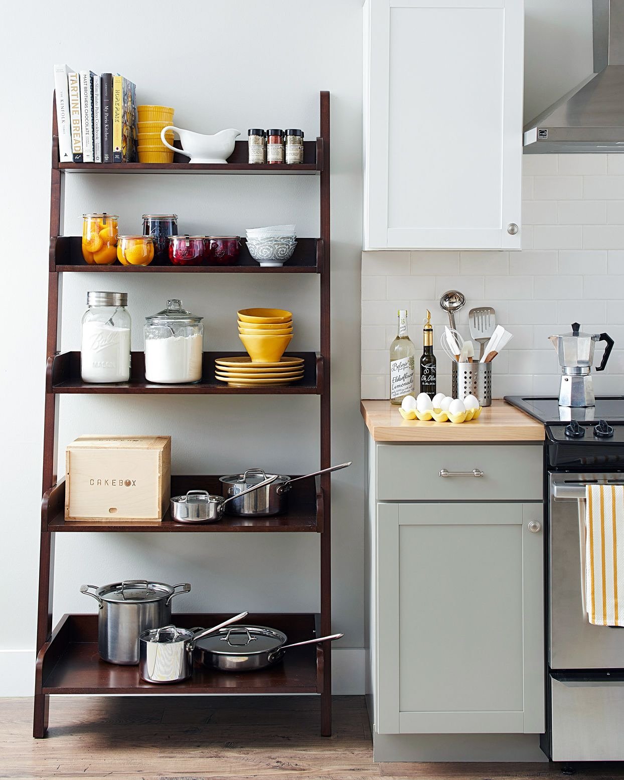 Dark brown, five-tier leaning ladder shelf organizing cookbooks, jars of dry goods, pots, and kitchen bowls next to a stove.