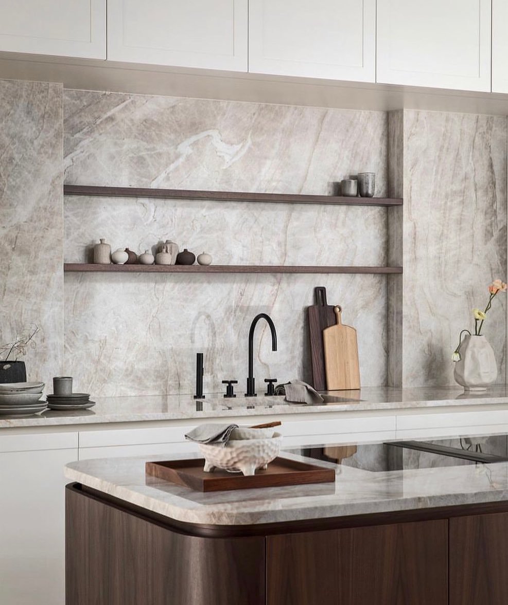 Modern kitchen detail showing a large, dramatic marble or quartzite backsplash with light veining, dark wood open shelves, and a contrasting dark walnut island front.