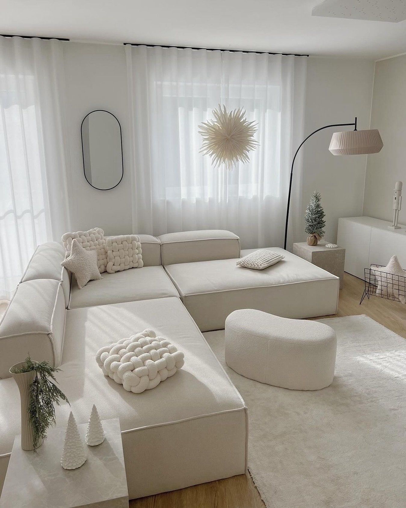 An ultra-modern, bright white and light beige living room featuring a large, low-profile modular sofa, a white sheepskin ottoman, and sheer curtains.