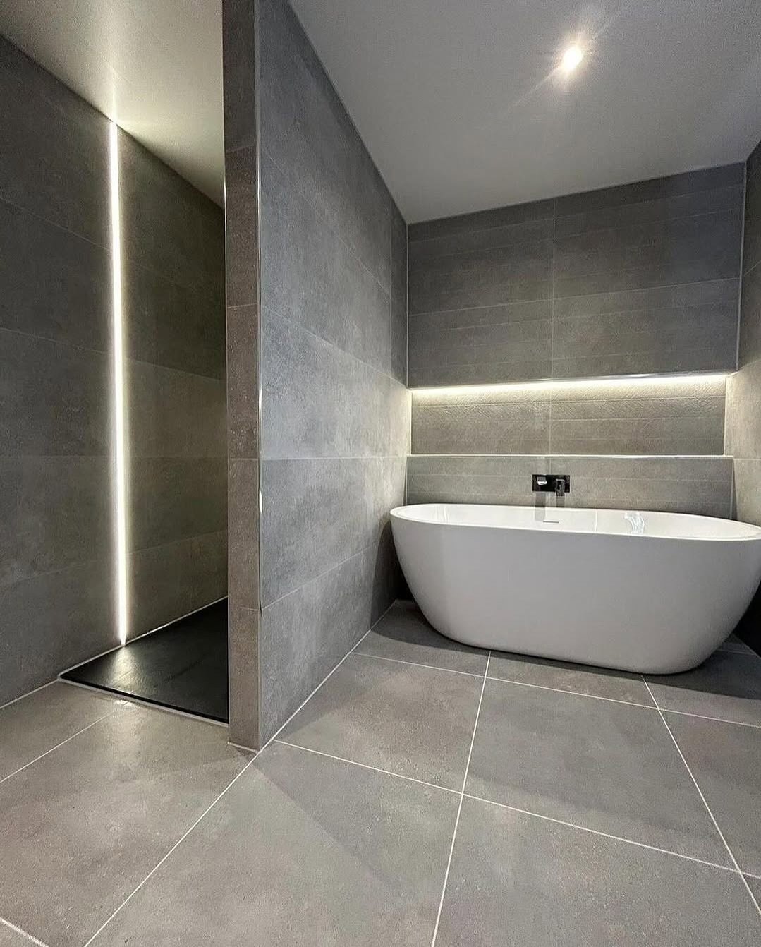 A minimalist, modern gray bathroom featuring a white standalone tub and large square concrete-look porcelain tiles on the floor and walls.