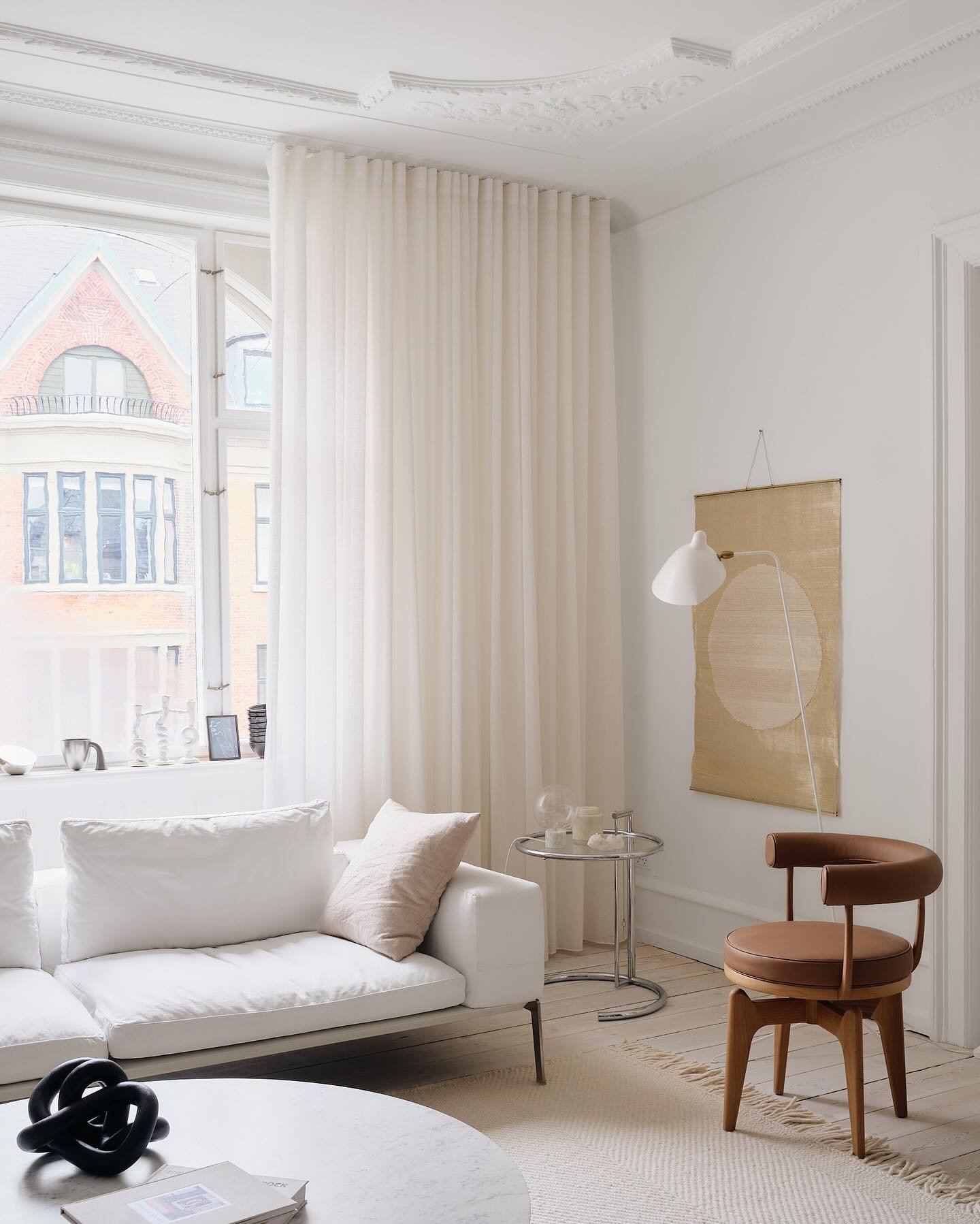 Floor-to-ceiling sheer white curtains covering a large window behind a modern white sofa and a leather wooden chair in a minimalist living room.