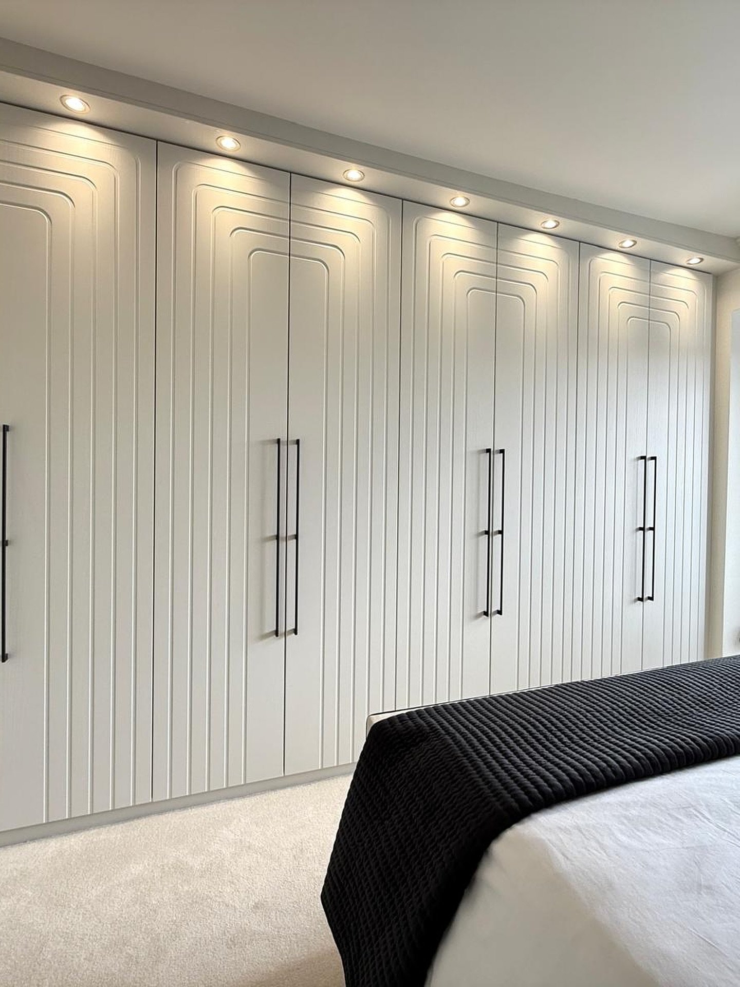 Modern white built-in wardrobe covering an entire wall, featuring vertical groove texture, subtle arched panels, black bar handles, and recessed lighting.