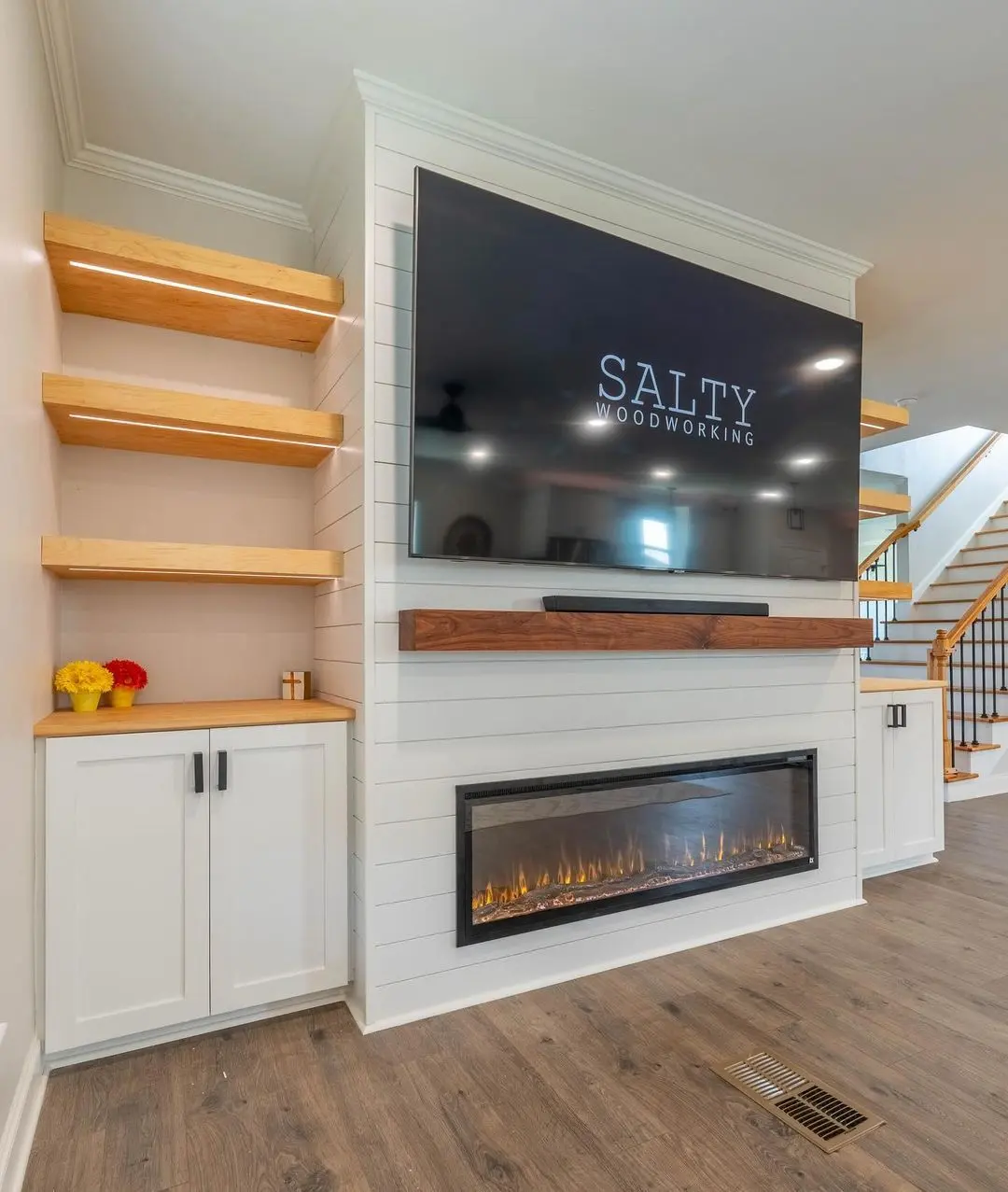 Transitional living space with an extra-long linear electric fireplace below a TV and walnut mantel, flanked by built-in white cabinets and LED-lit floating shelves.