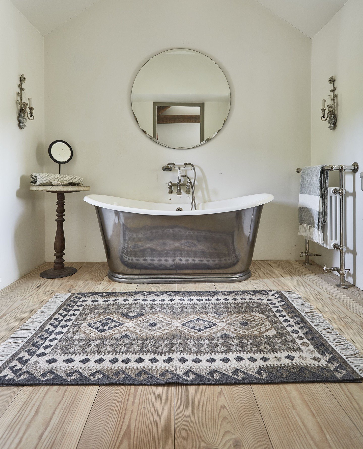 A rustic bathroom featuring light wood plank flooring, a polished metal freestanding clawfoot tub, a large round mirror, and a rectangular gray, white, and brown geometric patterned rug with tassels.