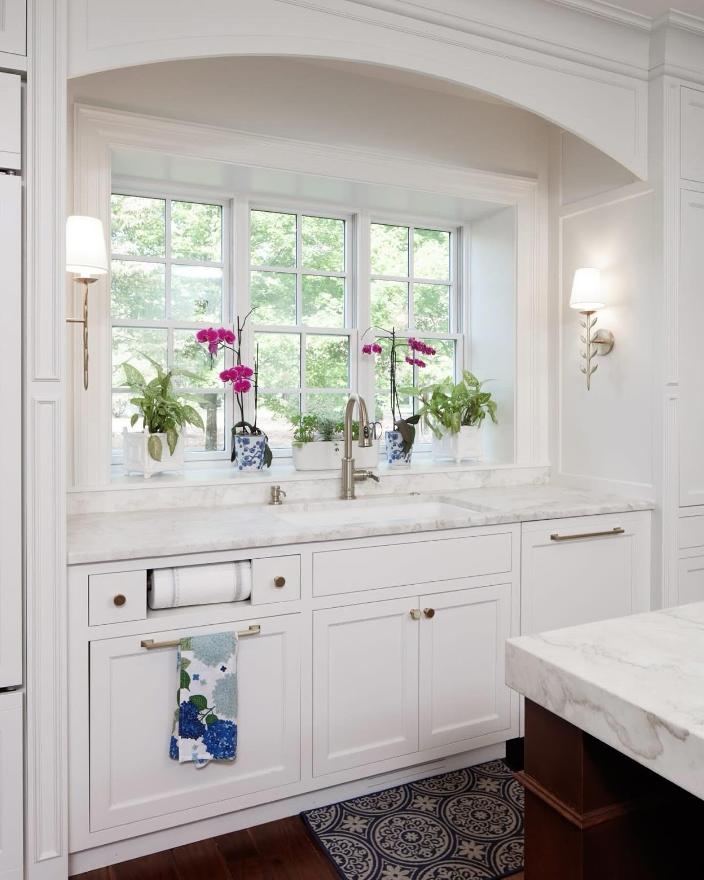 All-white classic kitchen featuring shaker cabinets, a marble countertop, and a white grid window set within a deep, arched trim above the sink. Orchids decorate the sill.