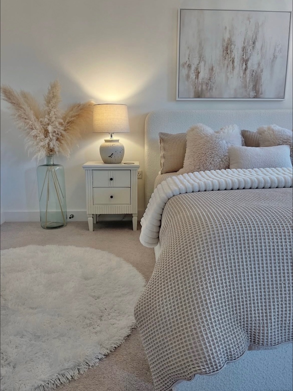 Close-up of a neutral bedroom featuring a white bouclé headboard, layered waffle and faux fur bedding, a white nightstand, and pampas grass decor in a tall glass vase.