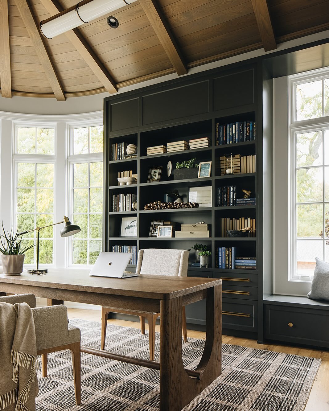 Luxurious home office with a dark charcoal built-in bookcase, a vaulted wood ceiling, large windows, and a heavy wooden desk over a plaid rug.