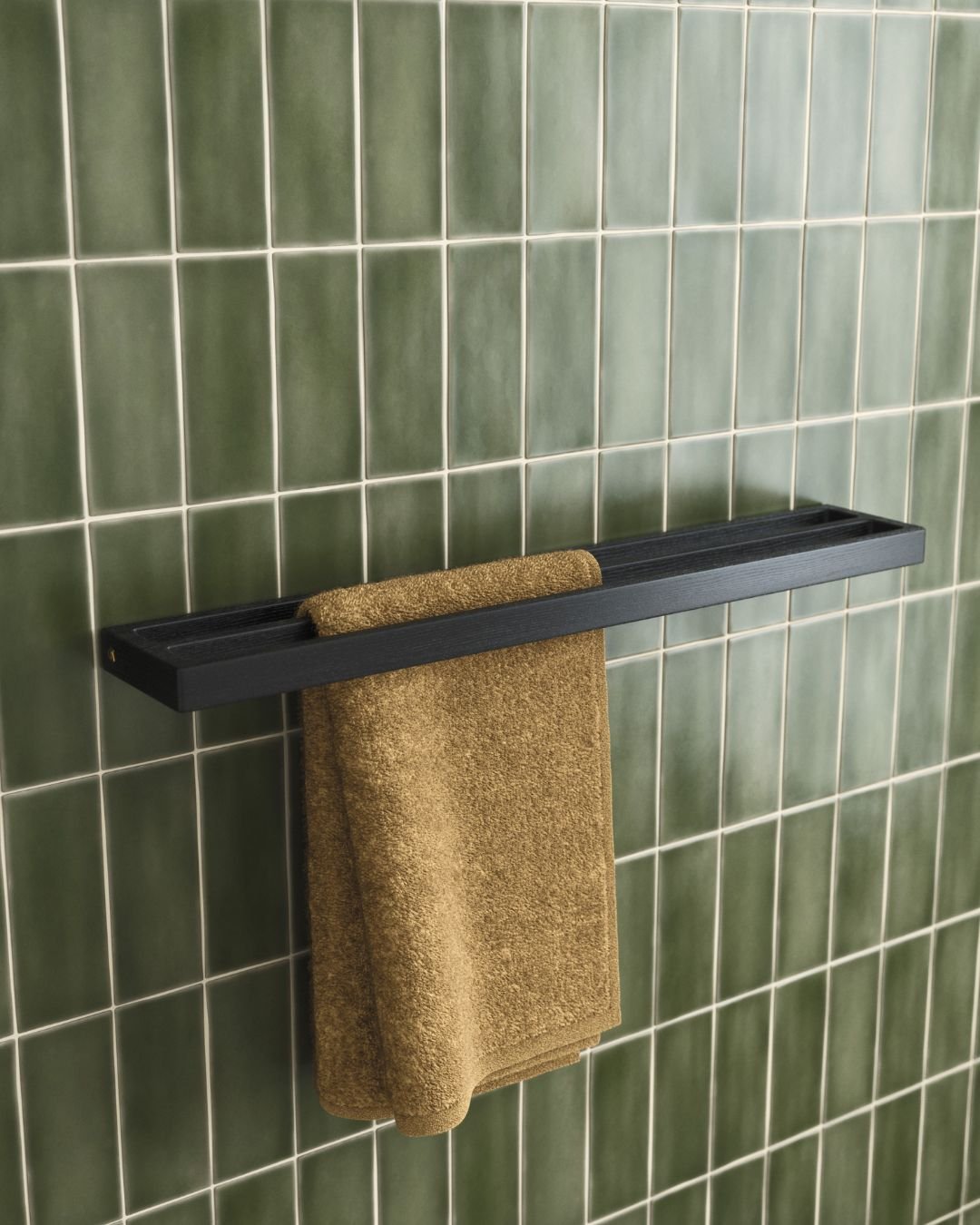 Dark wood or black floating shelf with an integrated towel hanging slot, mounted on a glossy emerald green subway tiled wall, holding a brown towel.
