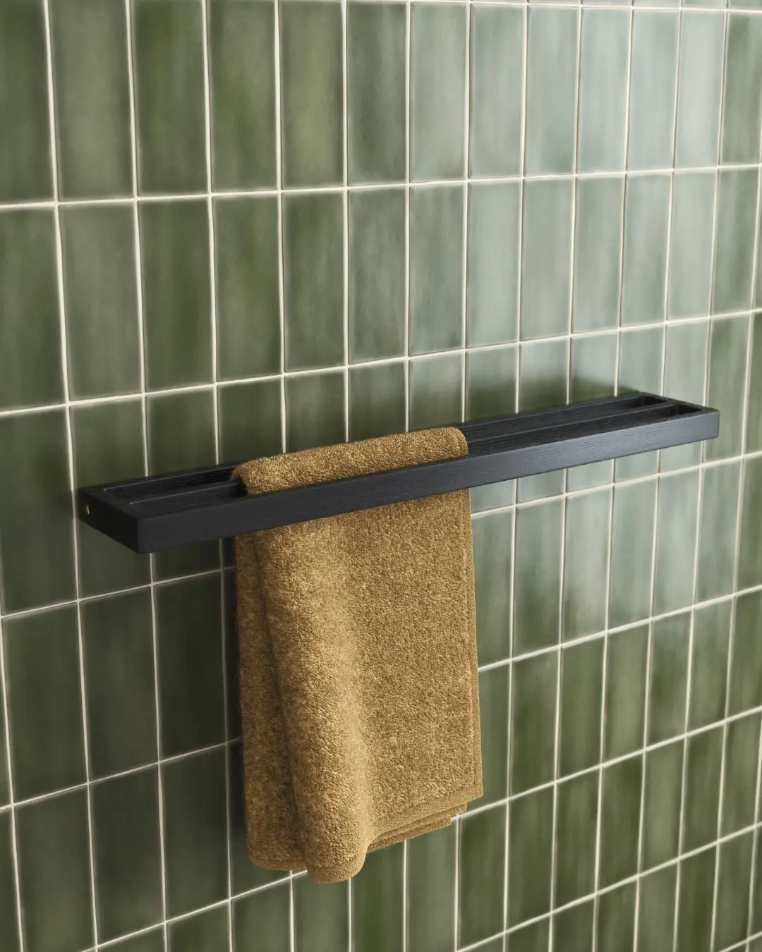 Dark wood or black floating shelf with an integrated towel hanging slot, mounted on a glossy emerald green subway tiled wall, holding a brown towel.