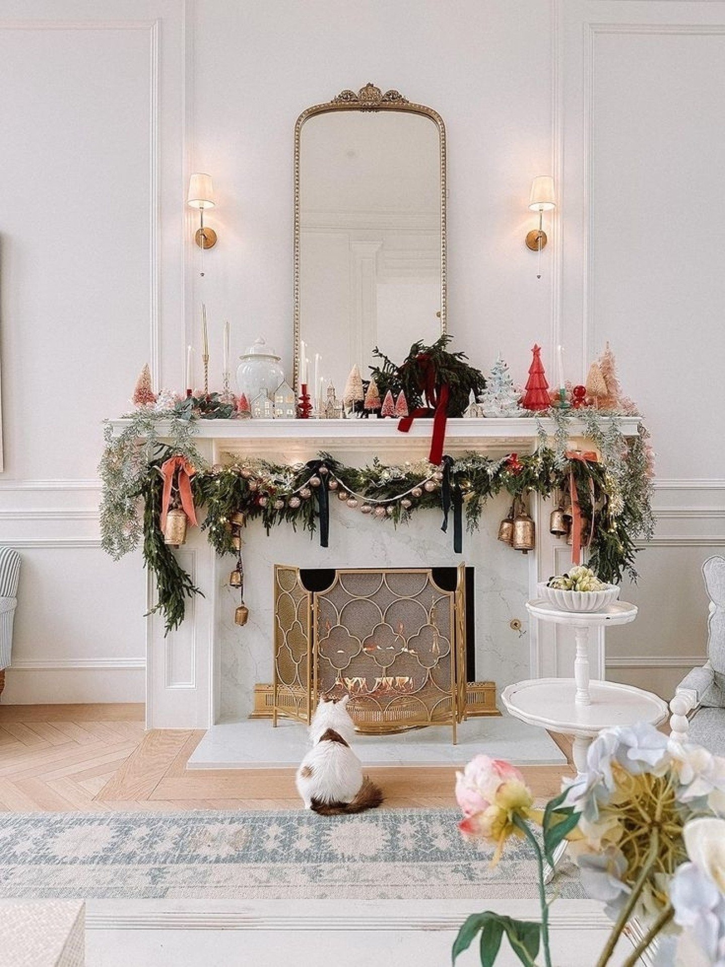 White mantel with a gold ornate mirror, decorated with layered garland, pink ornaments, deep red ribbons, and small vintage-style brush trees.