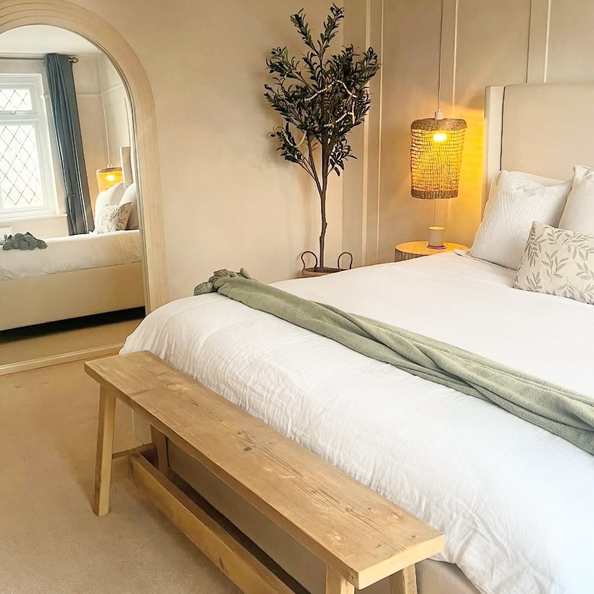 A bright bedroom with a bed covered in white linen, a large arched mirror, and a light-toned wooden trestle bench positioned in front of the bed.