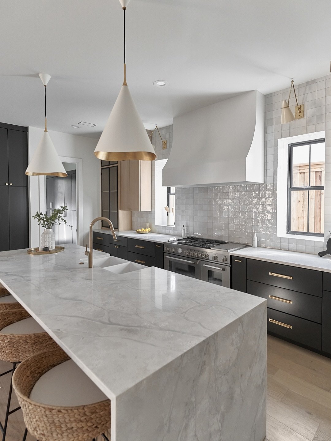 A bright kitchen featuring a black base cabinetry and a white marble waterfall island, accented by a white curved range hood and brass fixtures.
