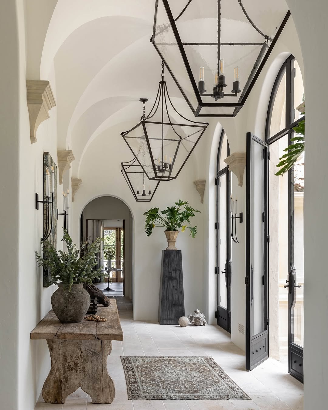 A long hallway with vaulted ceilings, large black metal lanterns, a rustic wood console table, and arched doorways.