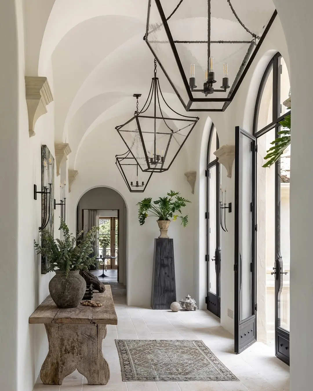 A long hallway with vaulted ceilings, large black metal lanterns, a rustic wood console table, and arched doorways.