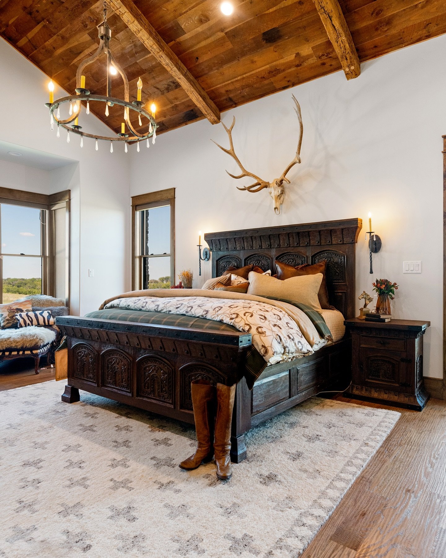 A luxurious Western bedroom with a massive carved dark wood bed, a wood-beamed ceiling, an antler mount, and an iron wagon wheel chandelier.