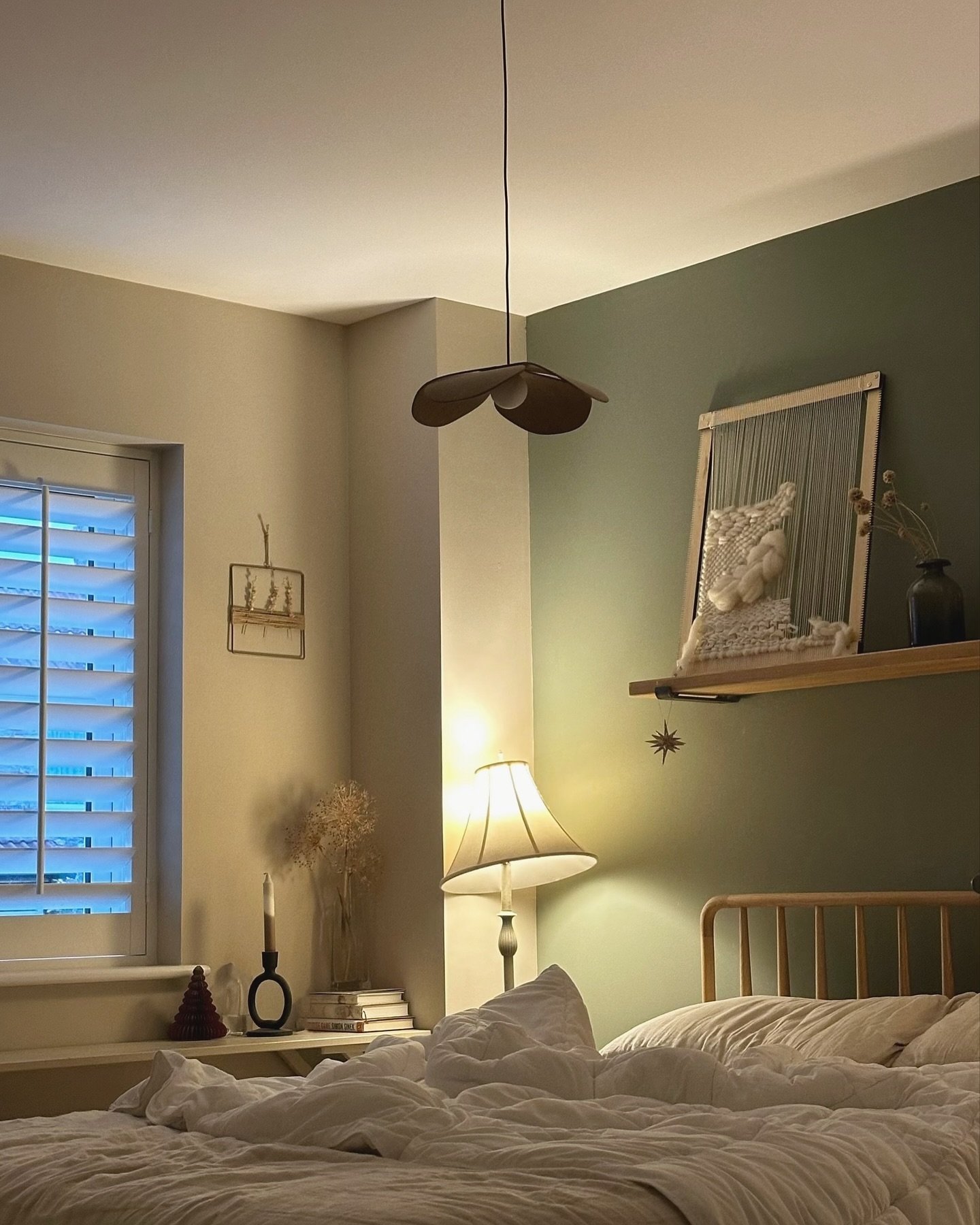 Dark and cozy bedroom with a deep sage green accent wall, warm lighting, and a woven tapestry on a wooden shelf.