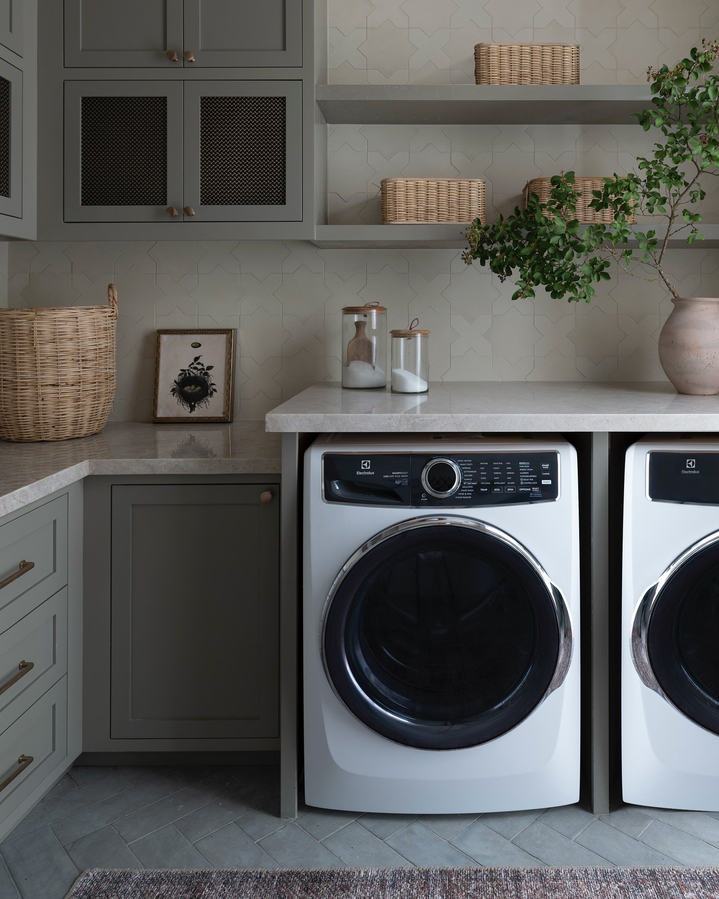 A close-up of a sophisticated laundry room featuring front-load washer and dryer beneath a light stone counter, sage-green cabinets, and a patterned tile backsplash.