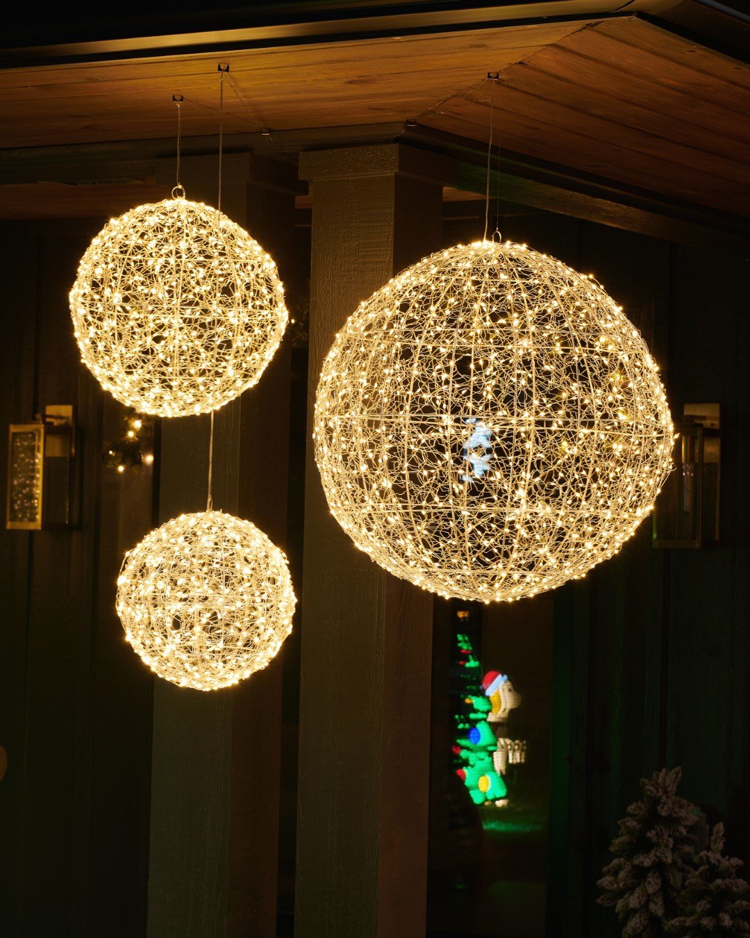 Three large, spherical light balls, made of woven wire and many small warm white LED lights, hang at different heights from a dark wooden porch ceiling.