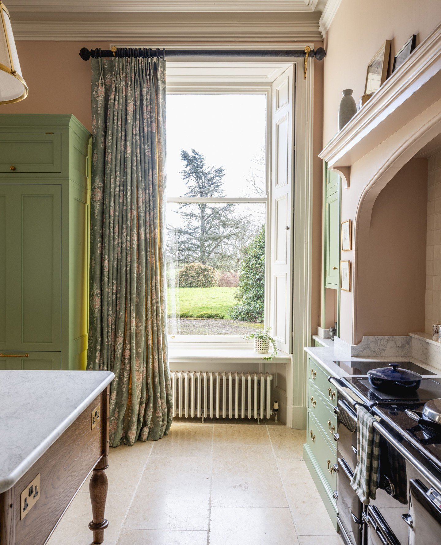 Classic kitchen with sage green cabinets, featuring a tall window with white internal shutters and a single, dramatic full-length curtain panel in a green and pink floral print.