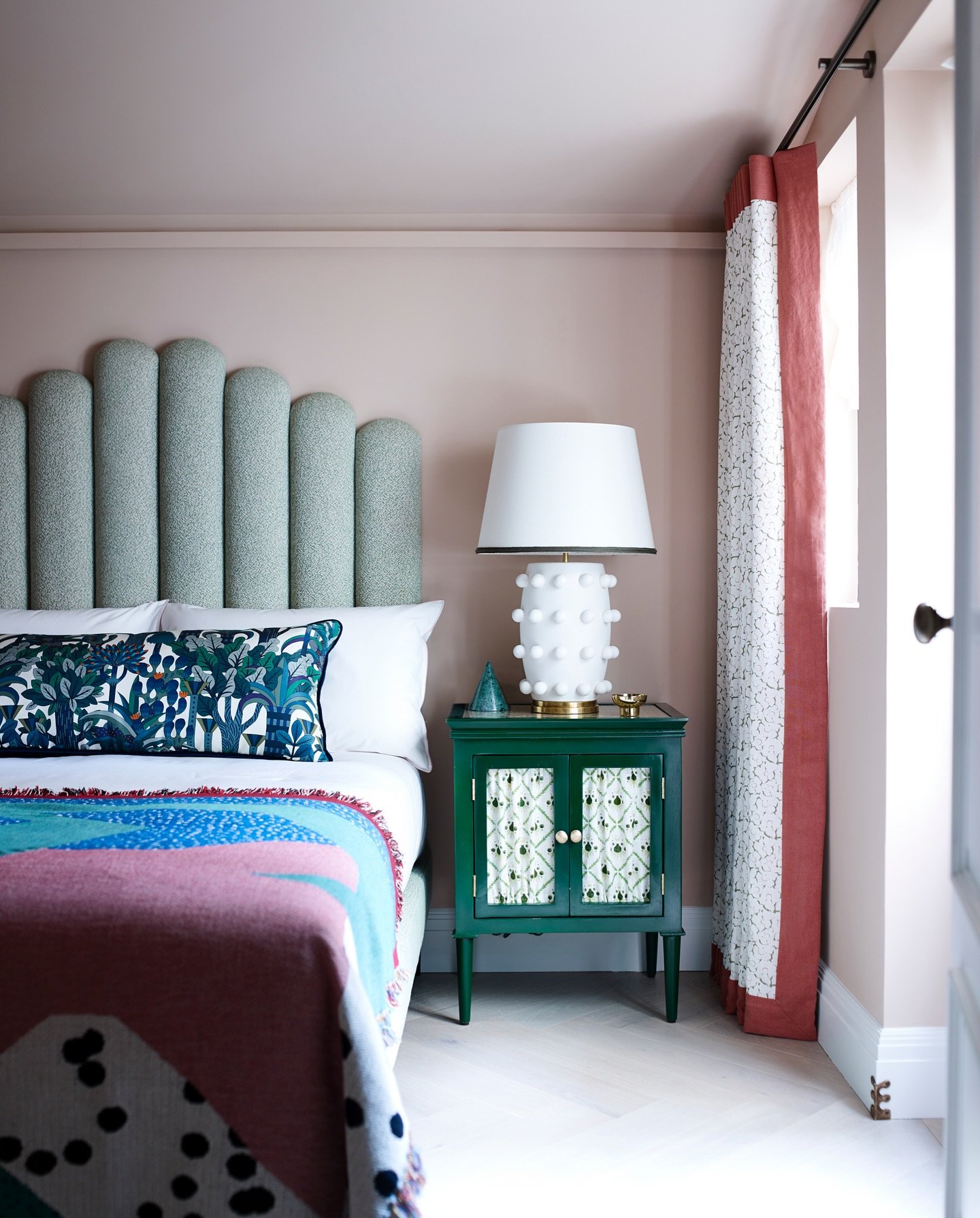 High headboard composed of several vertical, rounded, sage green upholstered flutes, positioned against a dusty pink wall in a maximalist bedroom.