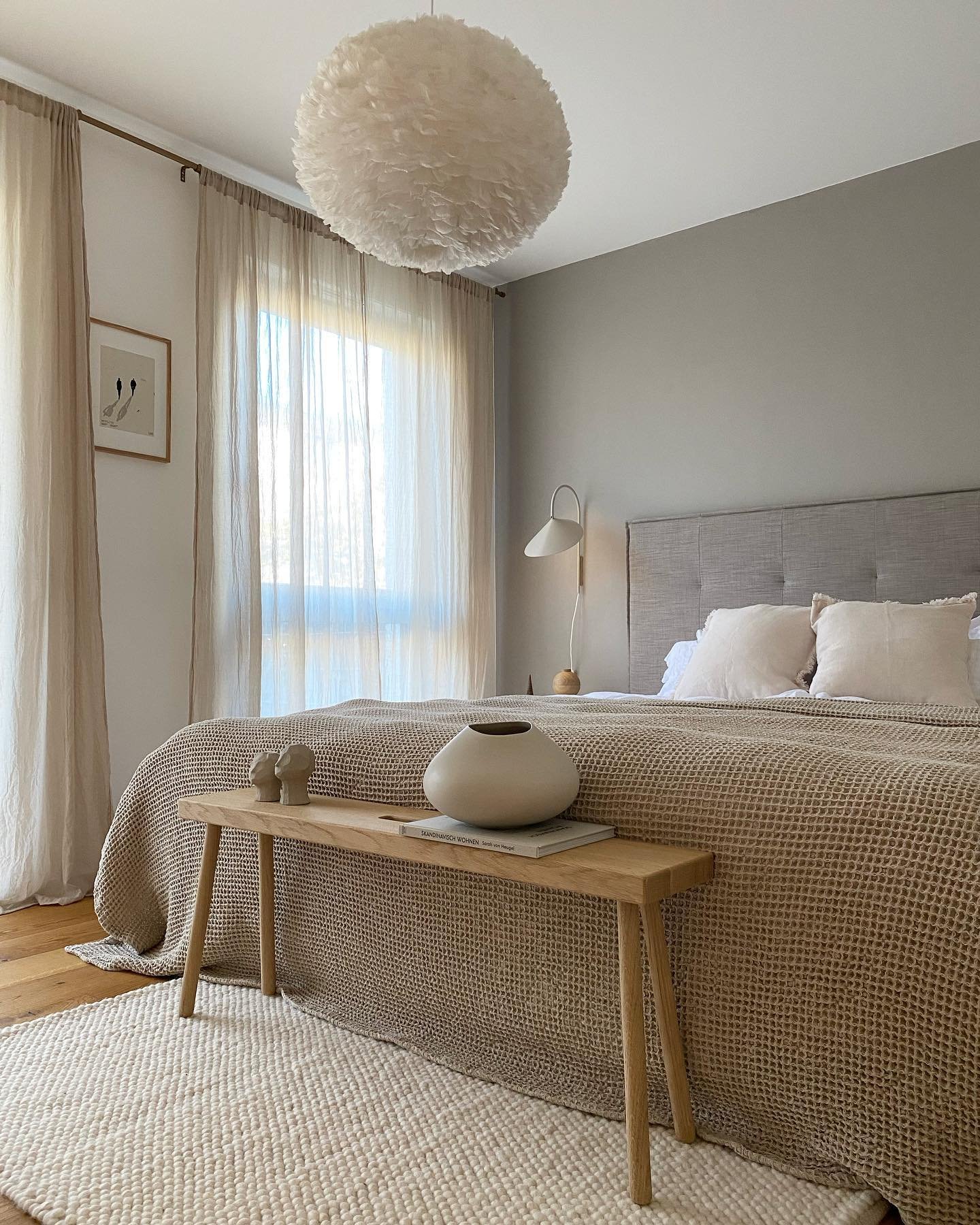 A cozy beige and gray Scandinavian-style bedroom featuring a light wooden bench at the foot of the bed, a chunky waffle-knit bedspread, and an oversized feather pendant light fixture.