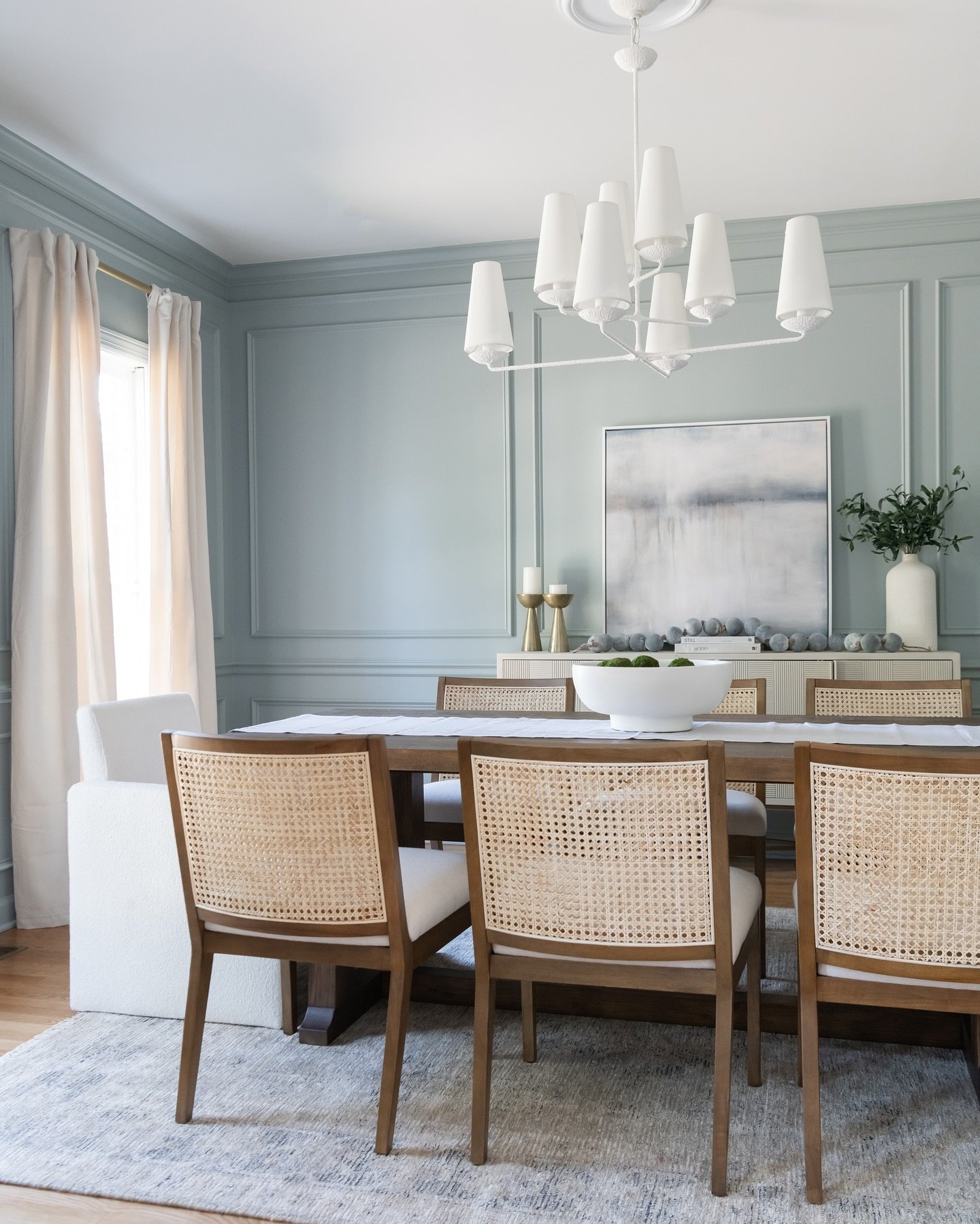 A bright dining room with dusty blue paneled walls, featuring a modern white chandelier, a long wooden table, and chairs with natural cane backs.