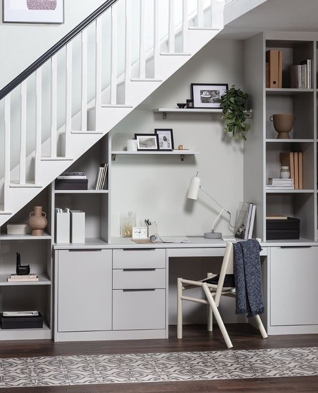 A light gray custom-built desk and storage cabinets beneath a white staircase.