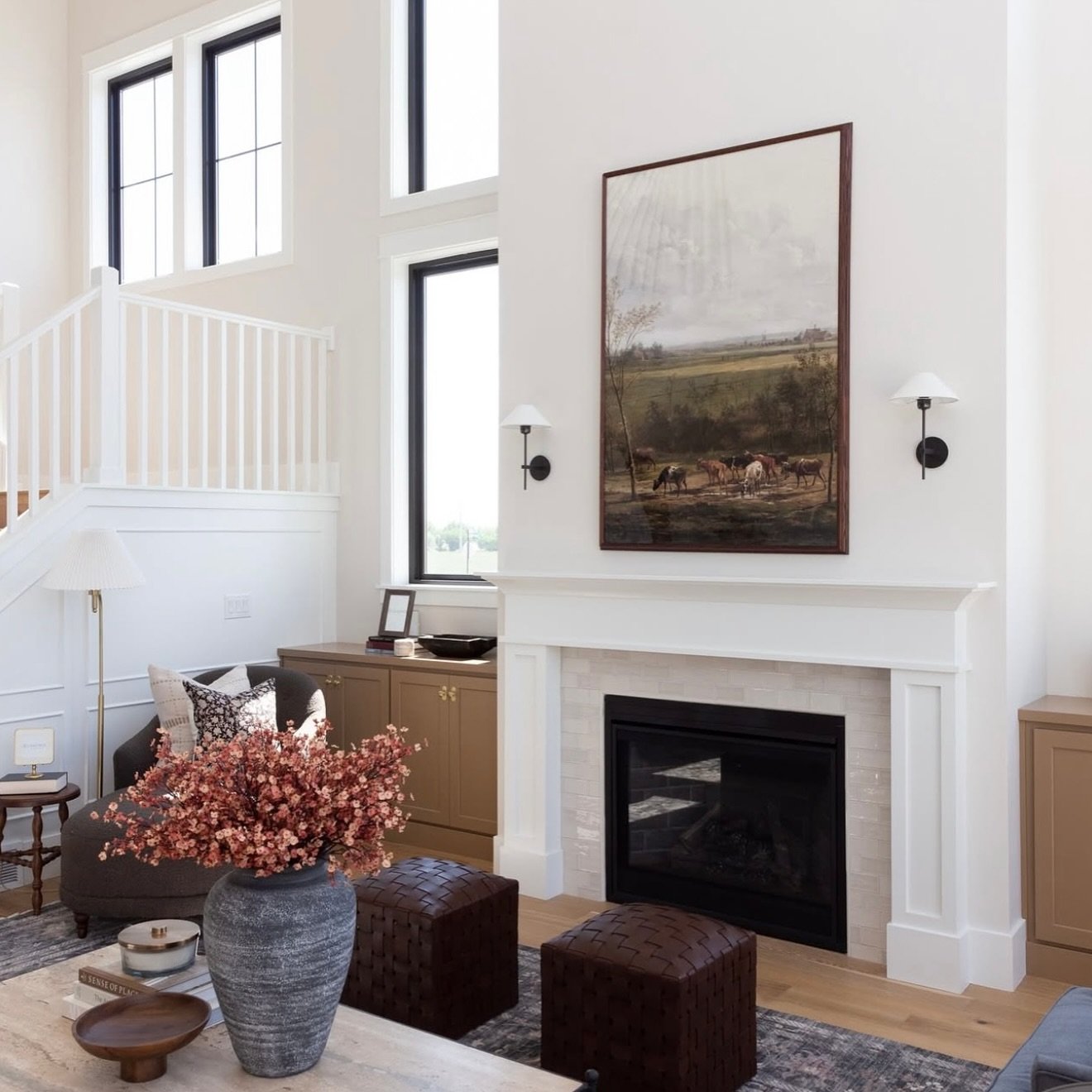 Two-story modern farmhouse living room with black-framed windows, a traditional white wood mantel, and tall framed pastoral artwork.
